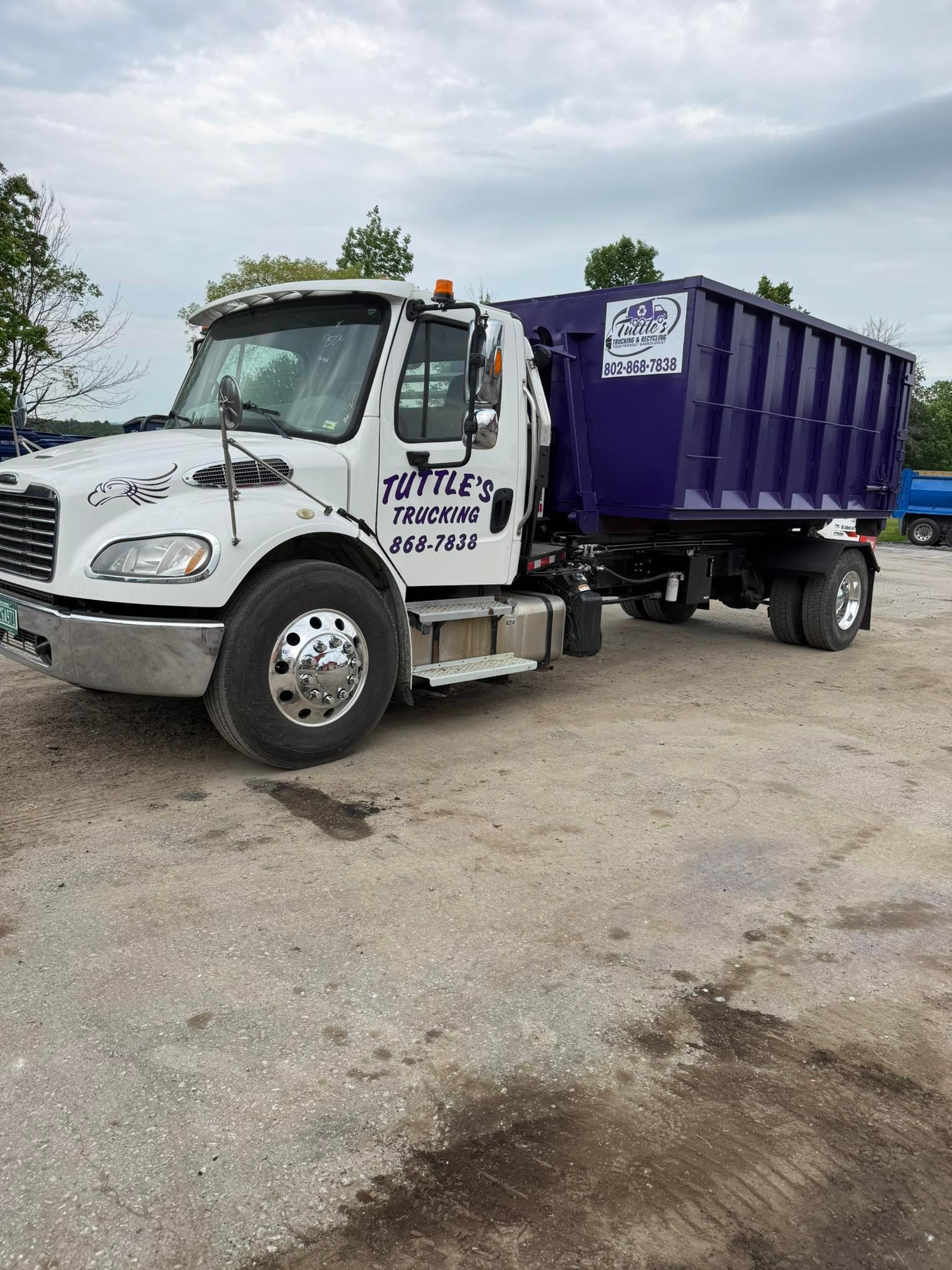 White dump truck with purple container; gravel lot, cloudy sky. 