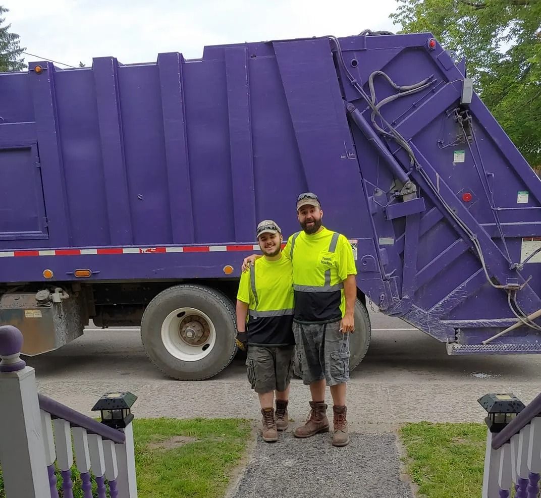 Two sanitation workers in neon shirts stand in front of a purple garbage truck.
