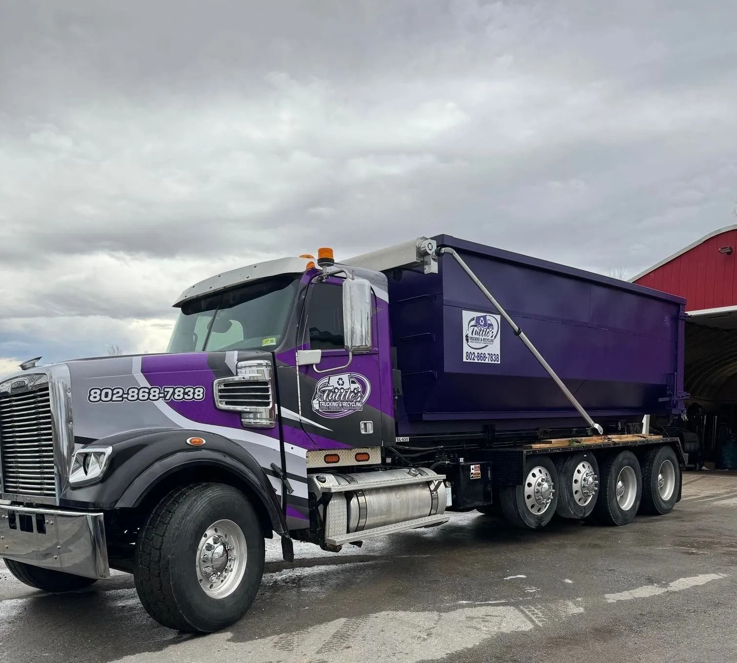 Purple and grey dump truck with logo, parked outside.