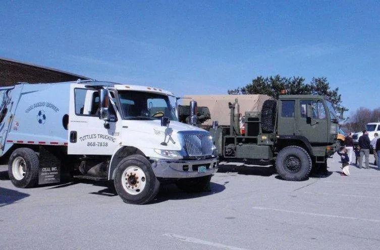 Two trucks parked outside on a sunny day. One is a garbage truck, the other a military vehicle.