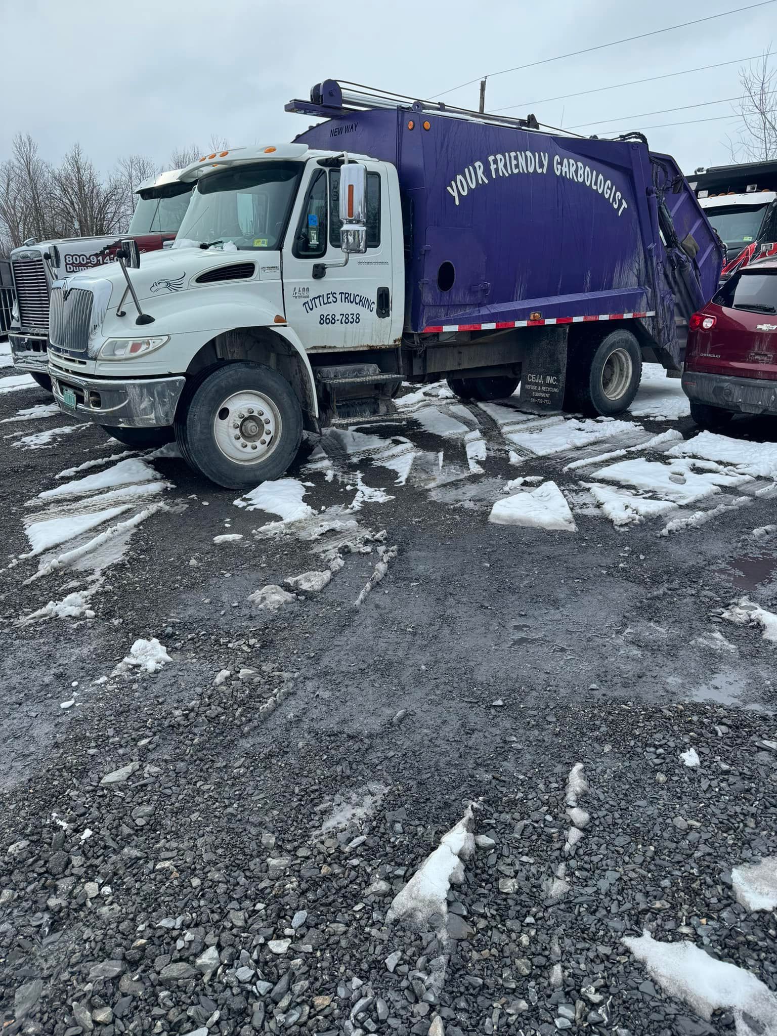 White and purple garbage truck parked on gravel in a snowy setting.