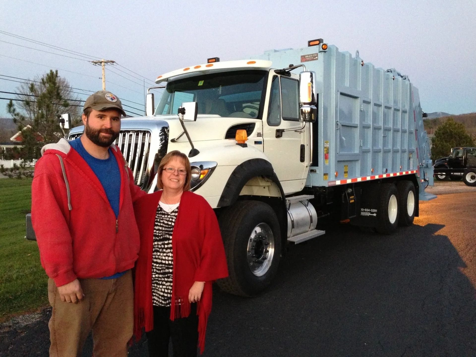 Man and woman pose next to a white garbage truck on a paved road.