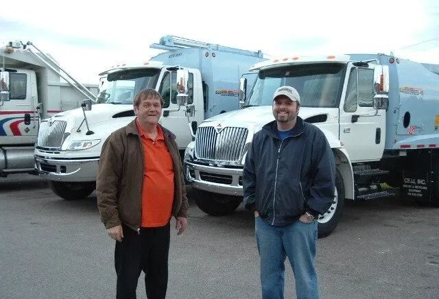 Two men standing in front of two white tanker trucks; one man wears an orange shirt and the other a blue jacket.