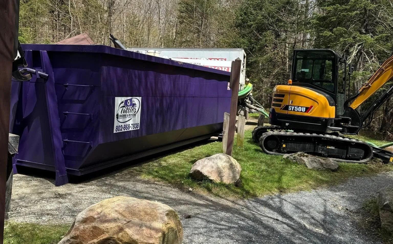 Purple dumpster next to a small excavator on grass with trees in the background.