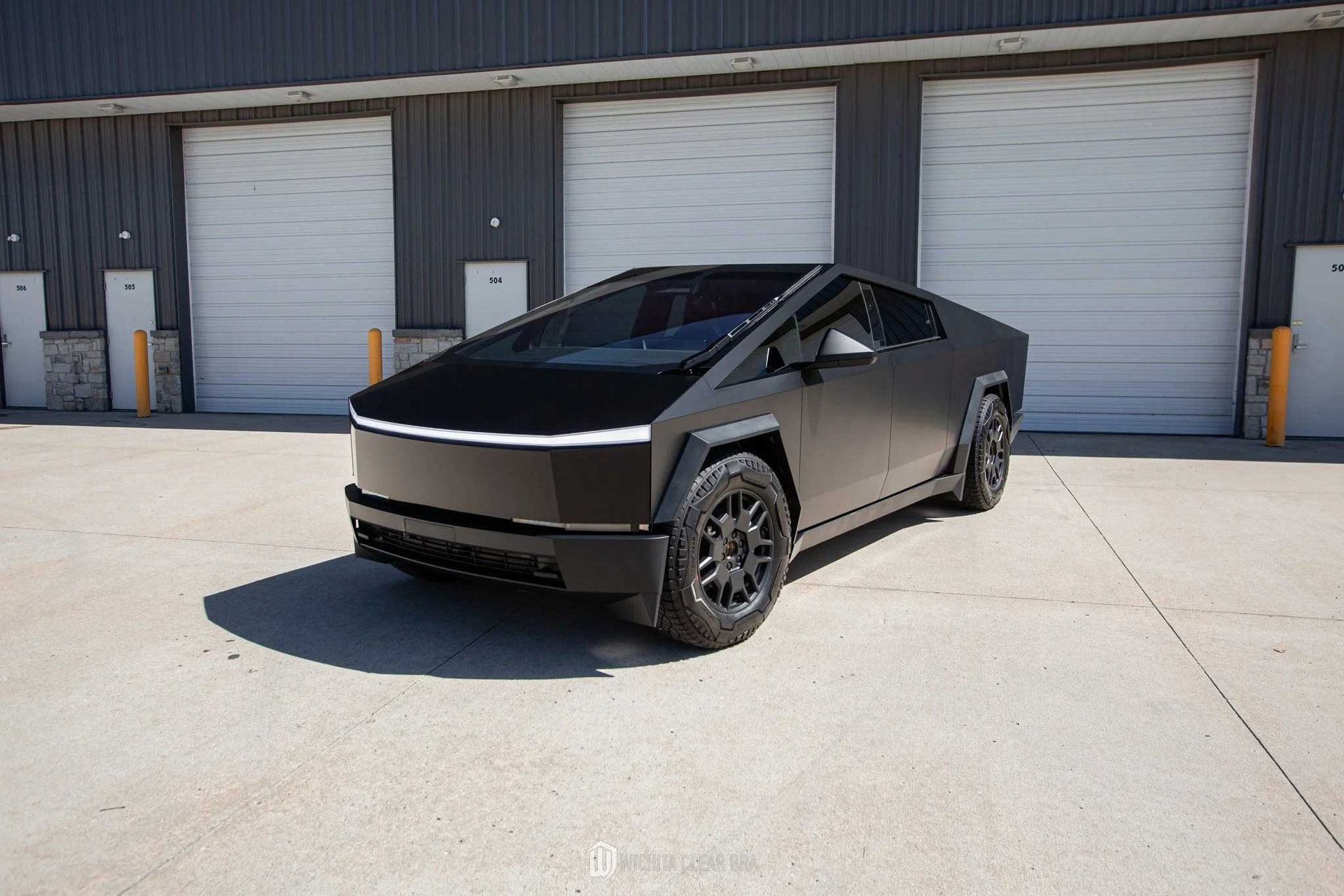 Black Tesla Cybertruck parked in front of a gray building with loading doors on a sunny day.