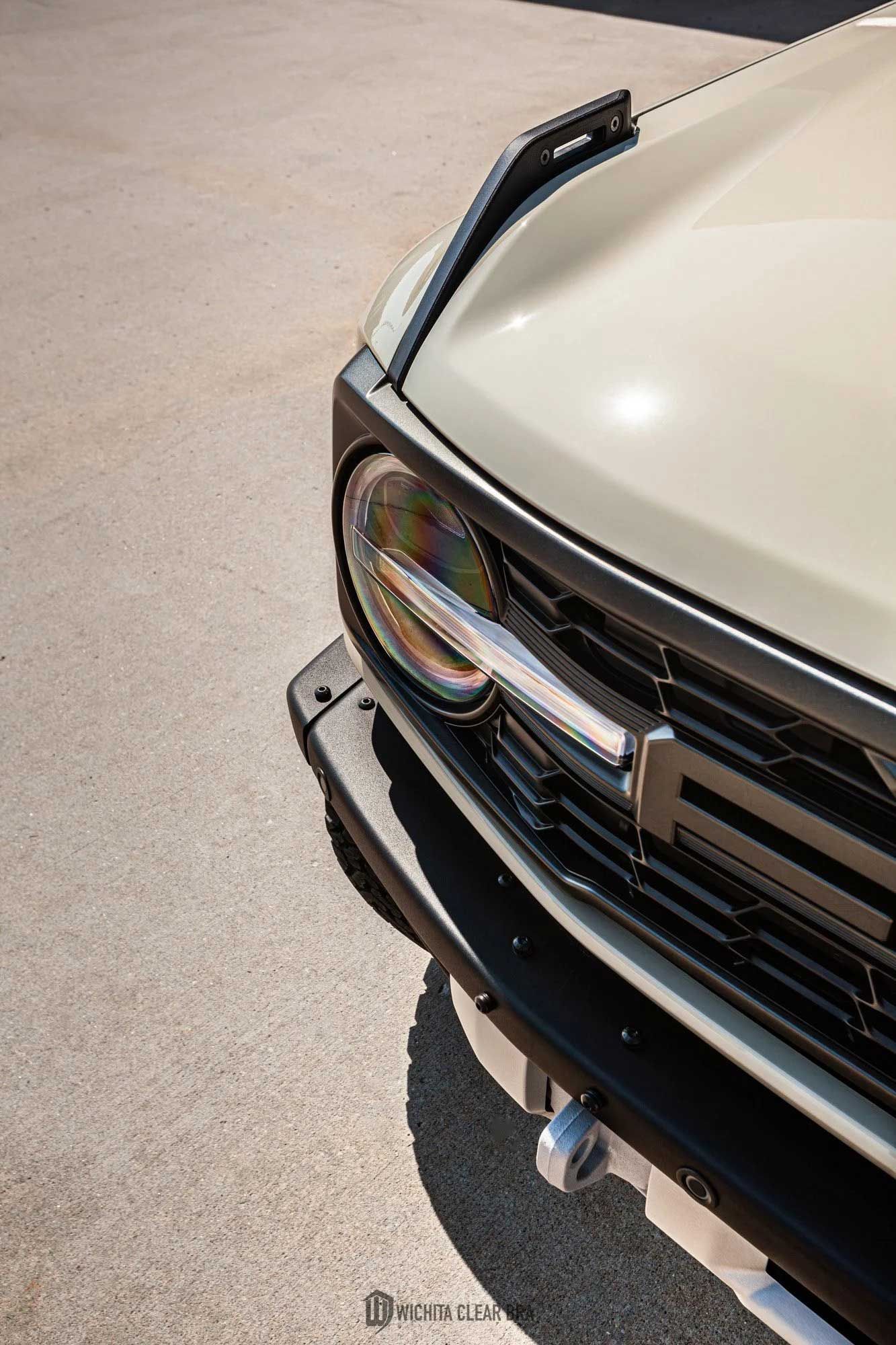 Close-up of a cream-colored Ford Bronco's front, showing the headlight, grill, and hood against a concrete background.
