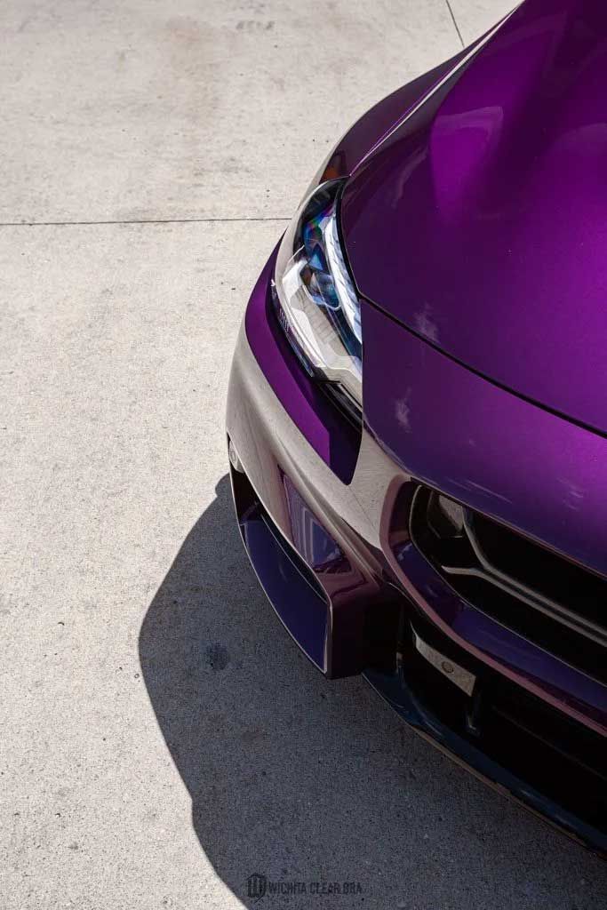 Purple car's front with bright headlights on concrete, casting a shadow.