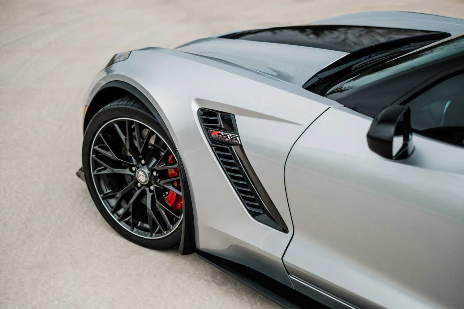 Silver sports car's front quarter panel; black wheel and side vent with ZR1 badge; red brake calipers.