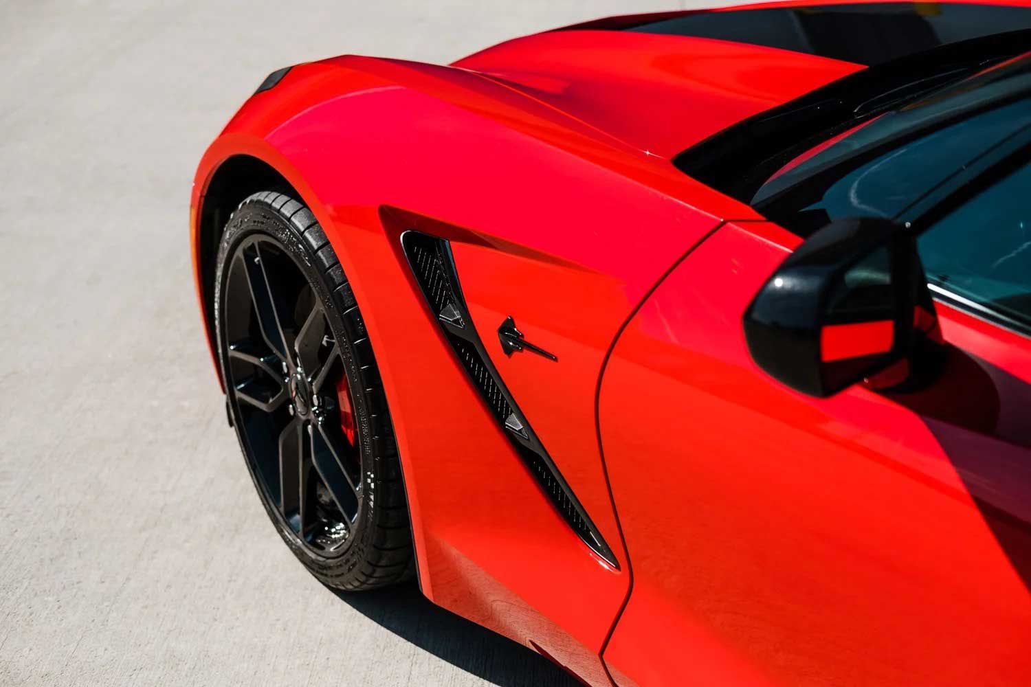 Bright red sports car's front quarter, with black accents on wheel, vent, and mirror, parked on concrete.