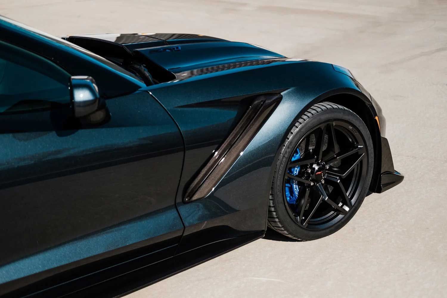 Dark blue sports car's front with a carbon fiber fender, black rim, and blue brake calipers.