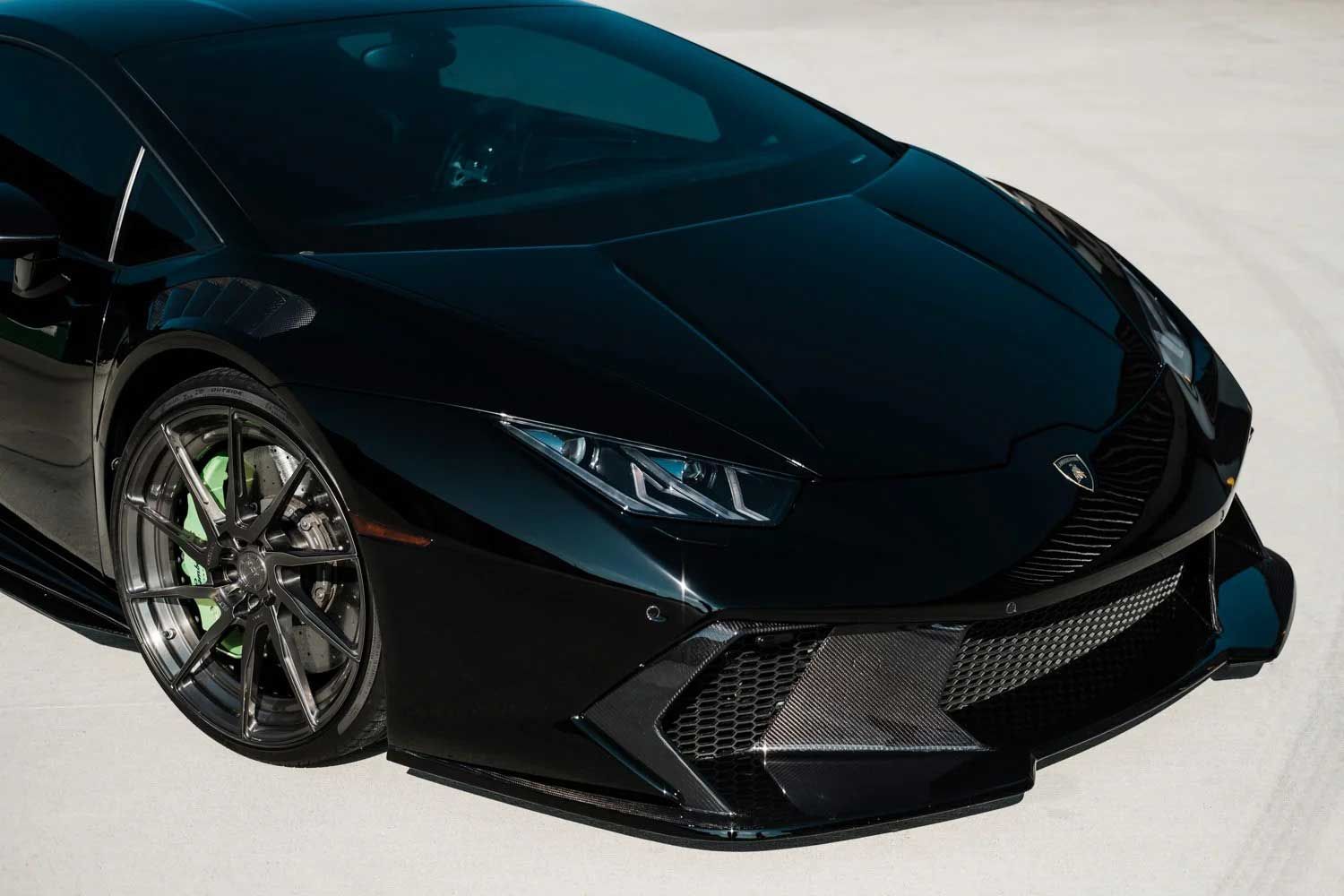 Black Lamborghini sports car on a white surface, showing the front end with a carbon fiber front splitter and green brake calipers.