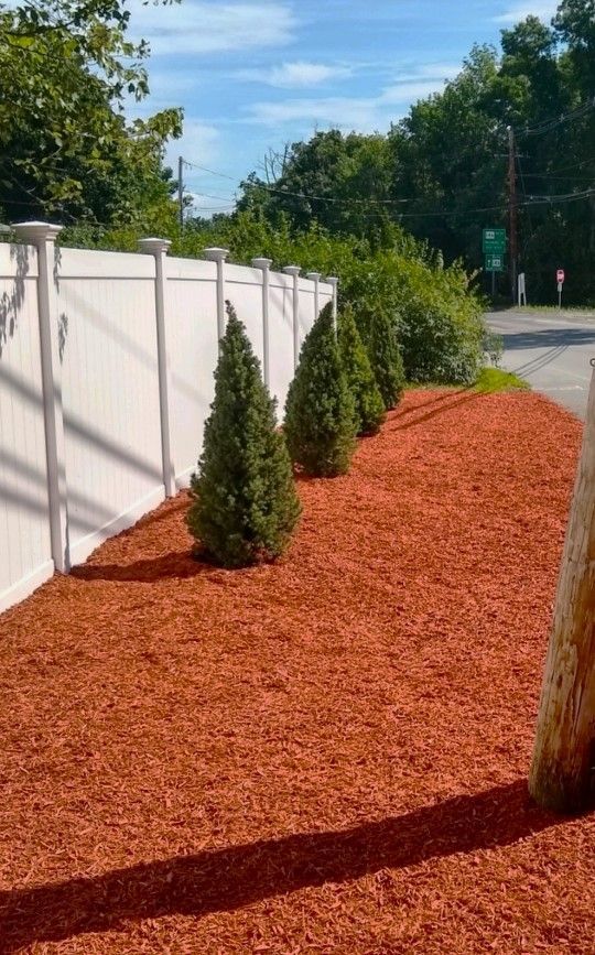 A white fence is surrounded by red mulch and trees.