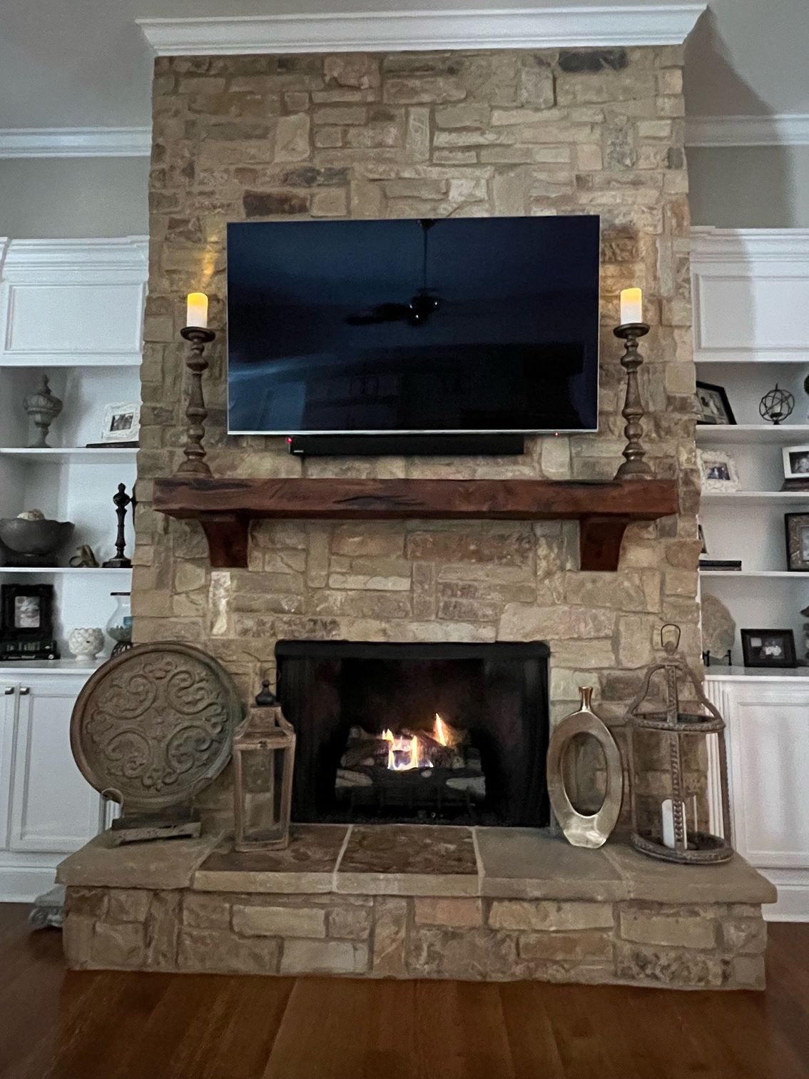 Stone fireplace with burning fire, TV, and wooden mantel. Decorative candles and lanterns surround.