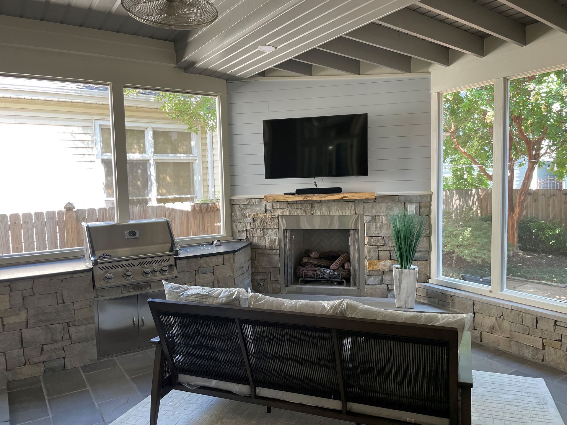 Outdoor living space with TV, fireplace, grill, and wicker sofa. Stone accents, windows, and trees are visible.