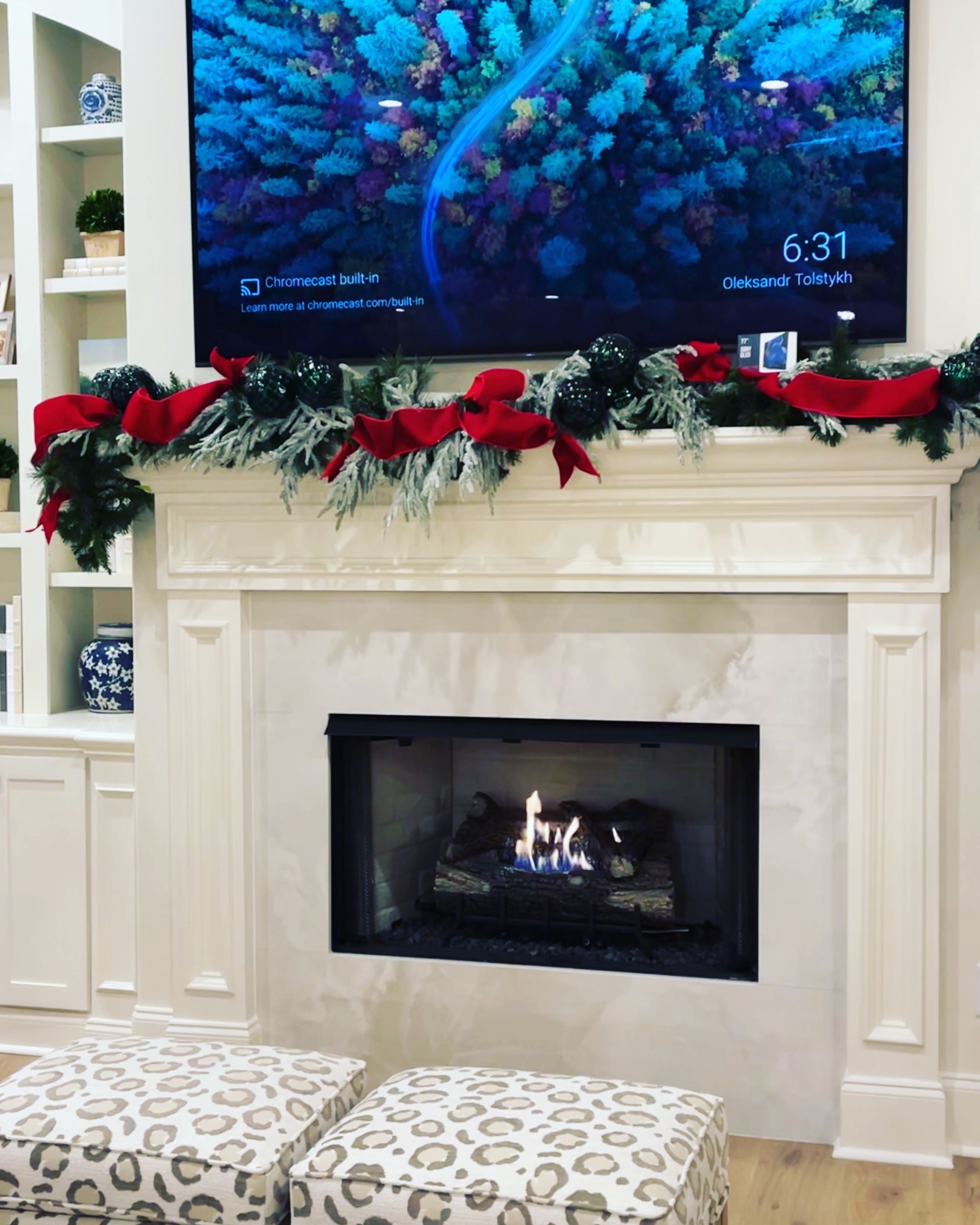 Fireplace decorated for Christmas with garland, red bows, and lit fire. A TV displays a nature scene.