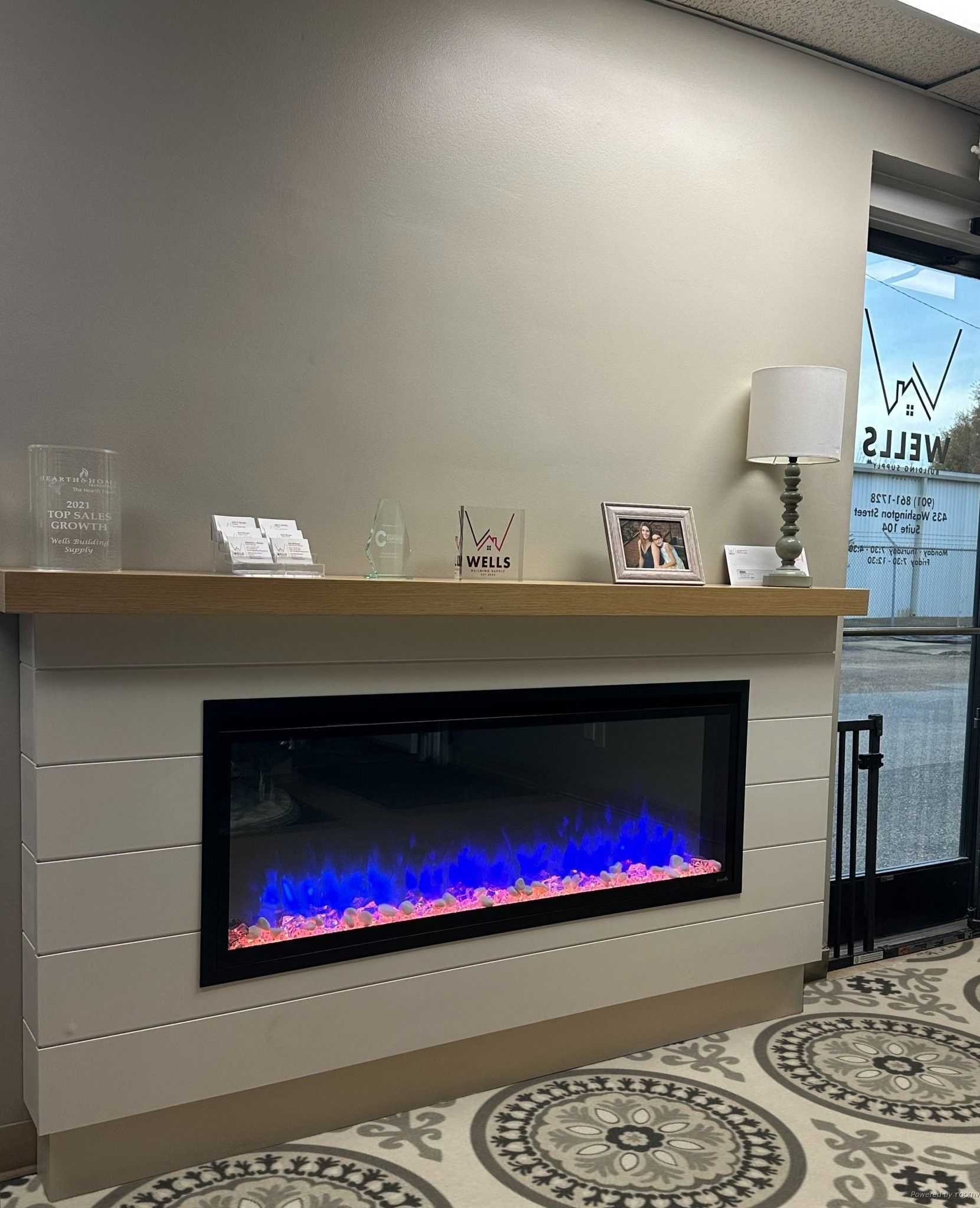 Electric fireplace built into wall, with blue flames, framed by light wood mantel and gray siding.