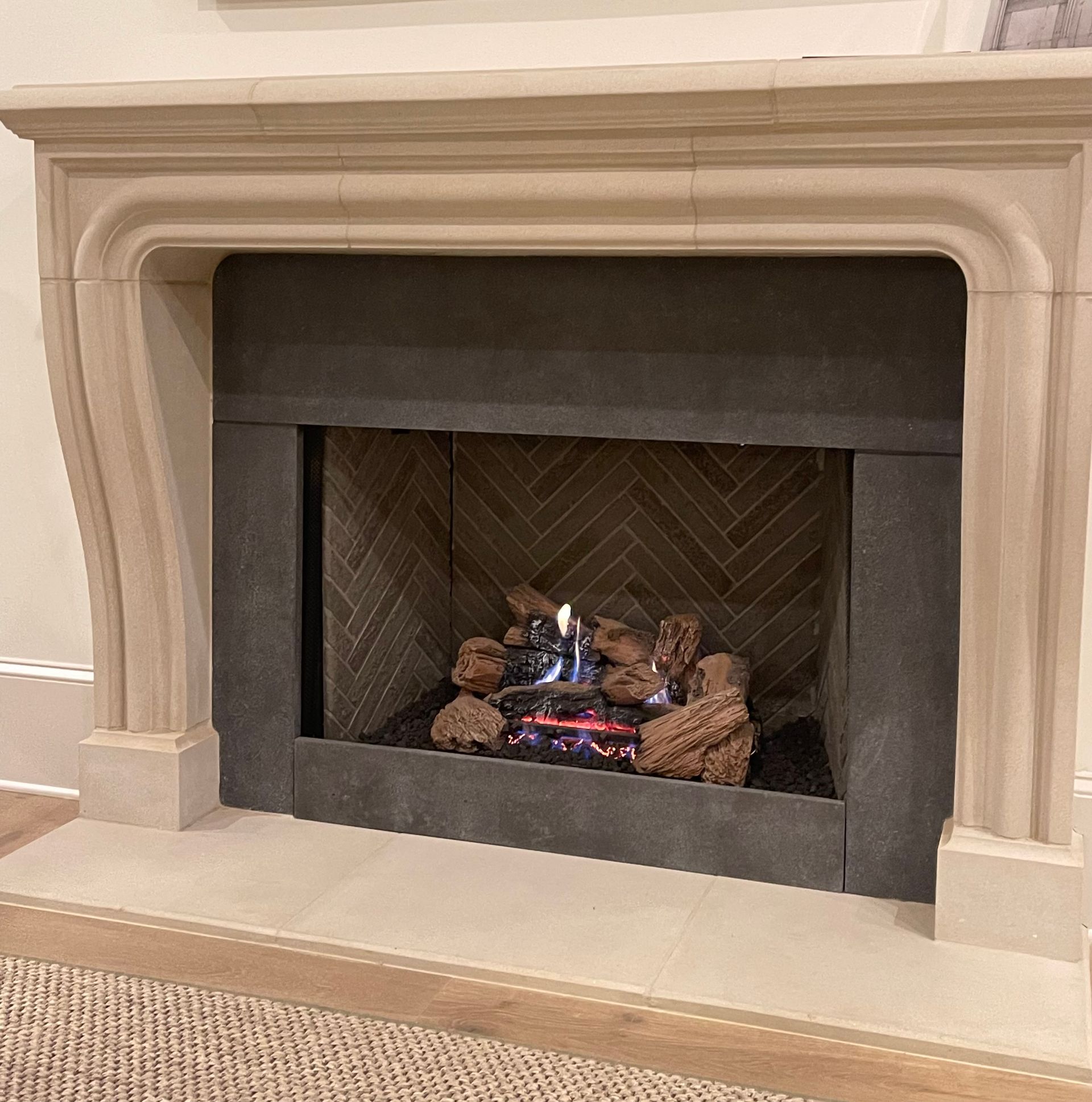 Fireplace with a stone mantel, lit gas fire, and herringbone patterned backing.