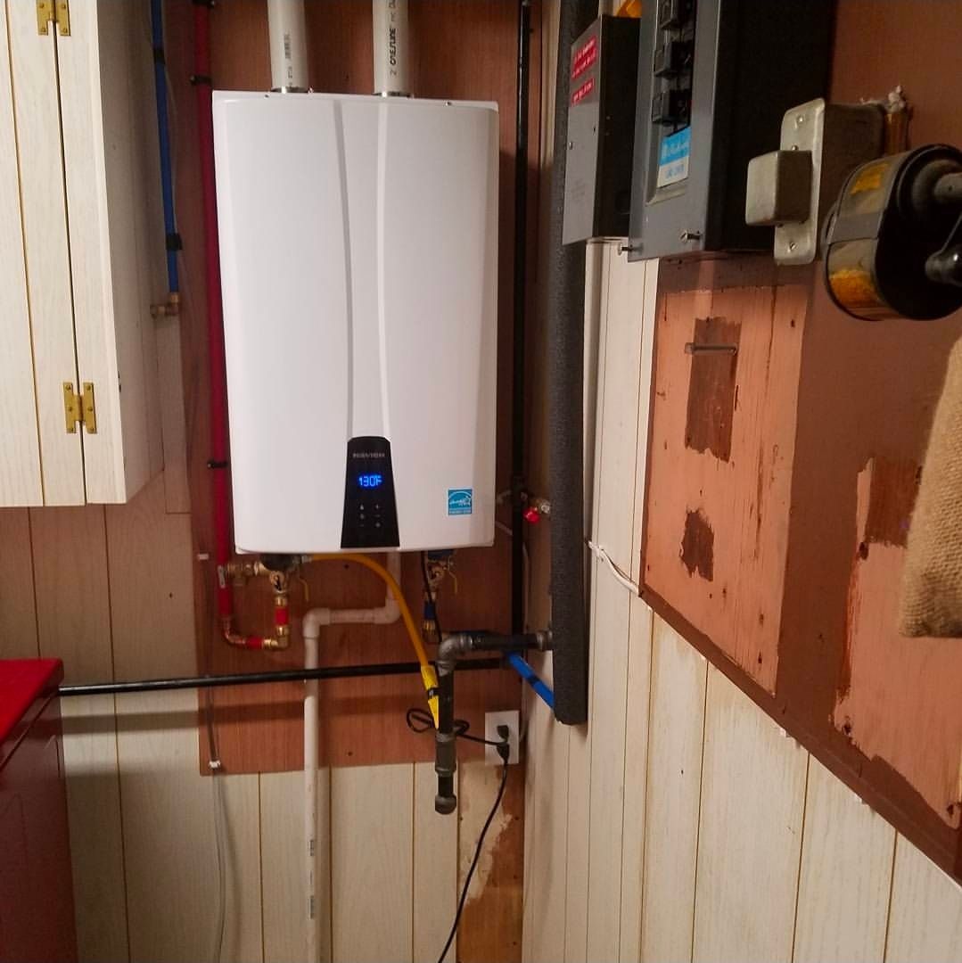 White tankless water heater on plywood-covered wall, with electrical panel and pipes.