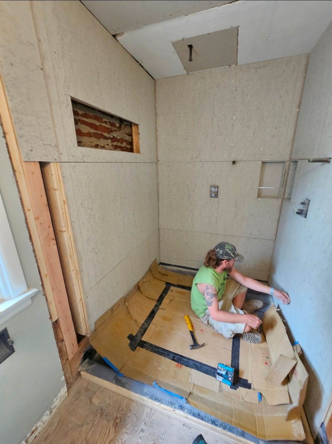 Person installing shower. Walls and floor are partially covered, some exposed brick. Hammer, tools visible.