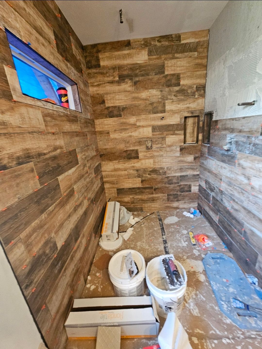 Bathroom under construction with wood-look tile on walls. Buckets, tools, and materials are on the floor.