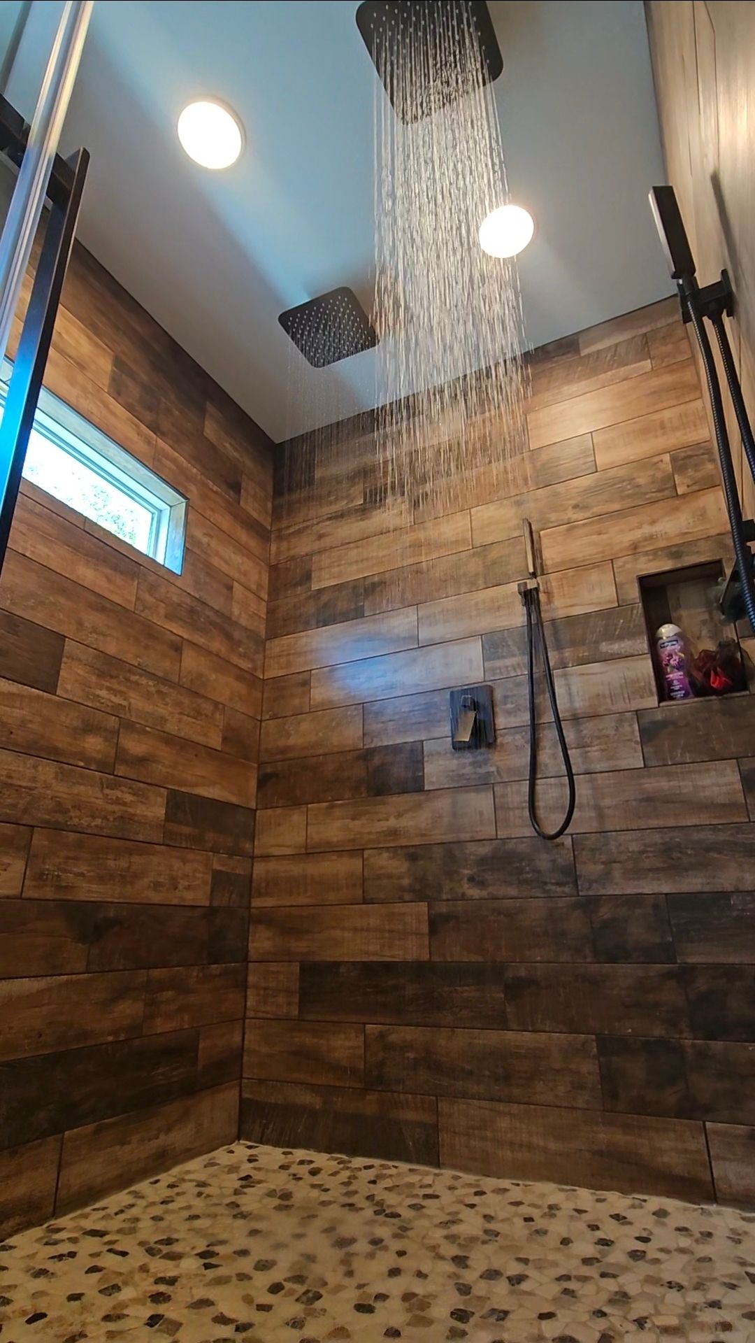 Shower with wood-look tile walls, pebble floor, and rain showerhead. Water streams down.