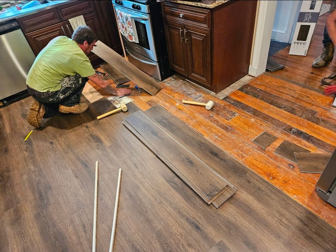 A person installing wood-look flooring in a kitchen. They are using tools on boards, near appliances and cabinets.