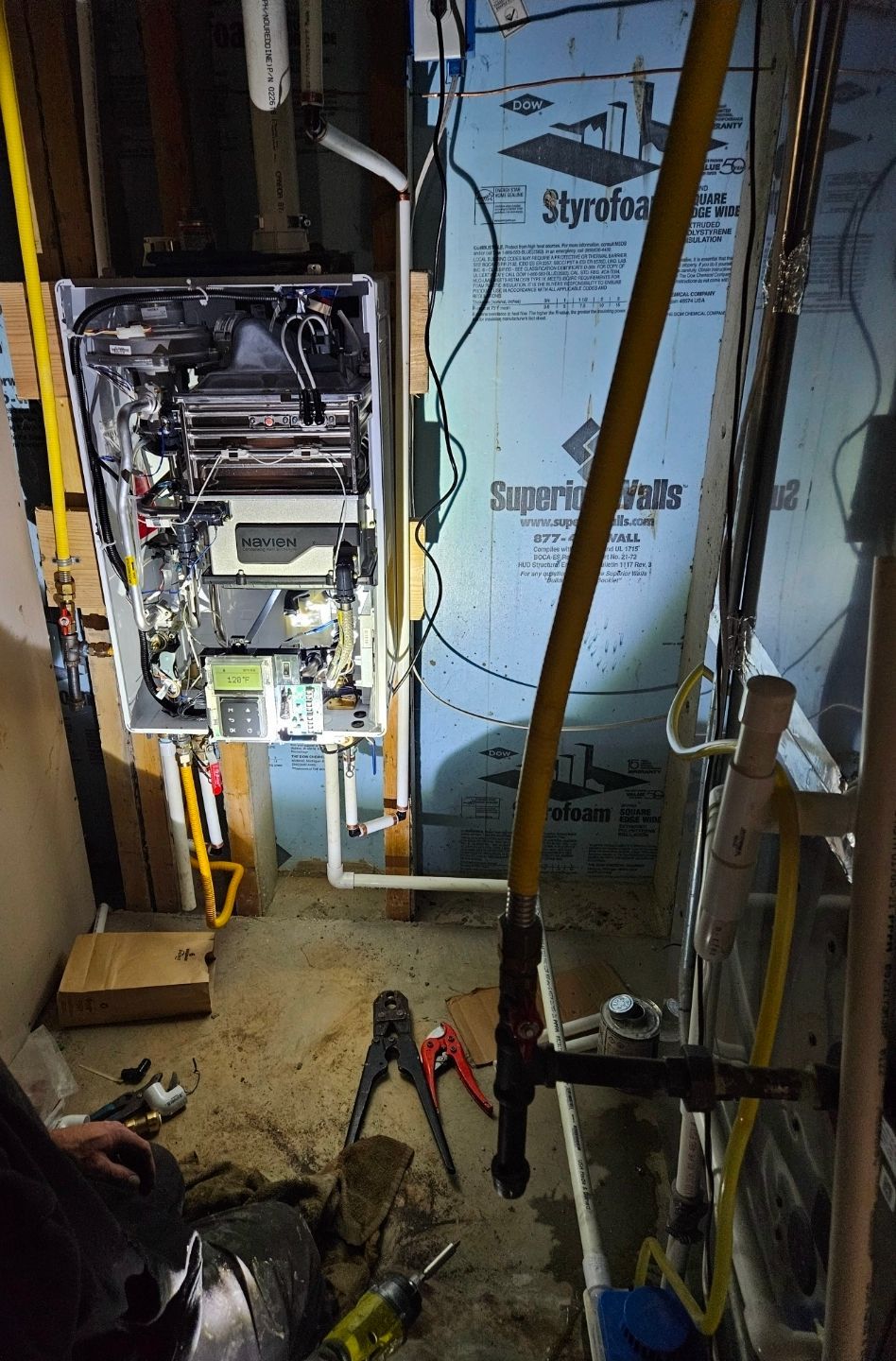 A person working on a gas boiler in a basement. Yellow gas lines and tools are visible.