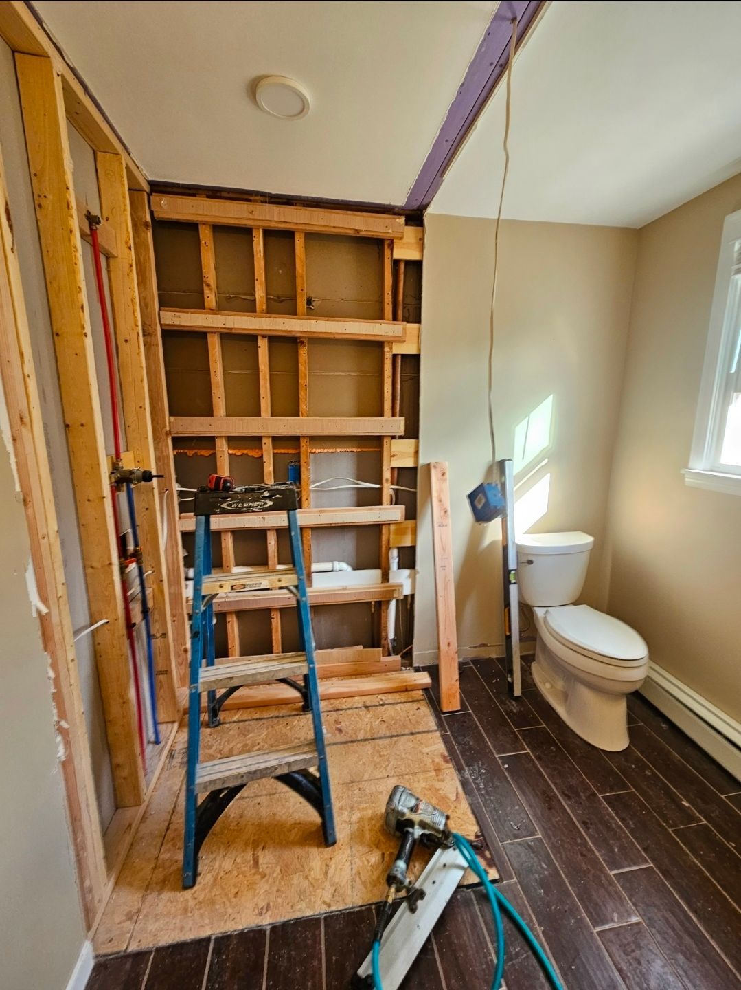 Bathroom undergoing renovation; exposed wall studs, toilet, ladder, and tools. Brown flooring and tan walls.