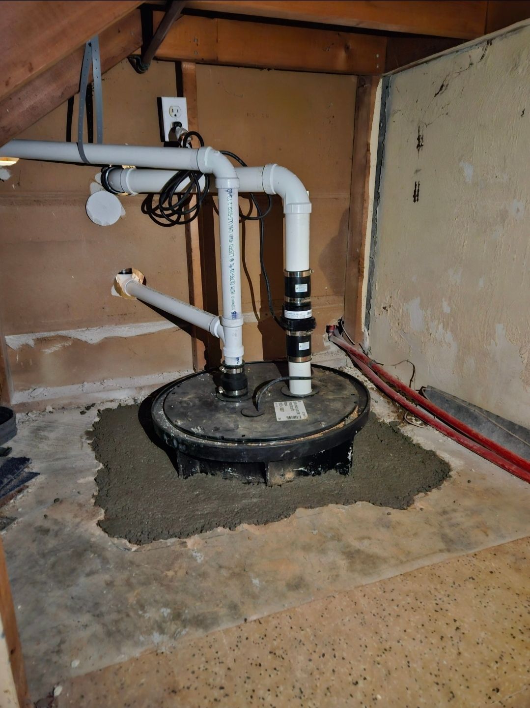 Sump pump installation in a basement. White pipes connect to the black pump on a concrete base.