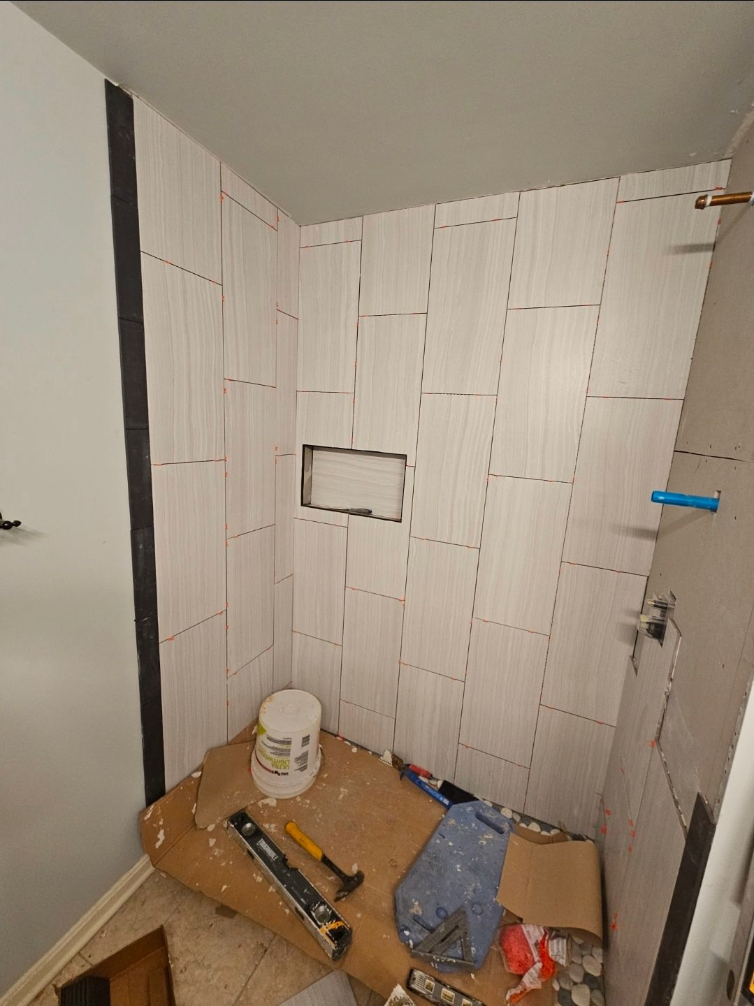 Bathroom remodel in progress; tiled shower walls with a niche. White tiles, mortar bucket, tools on the floor.