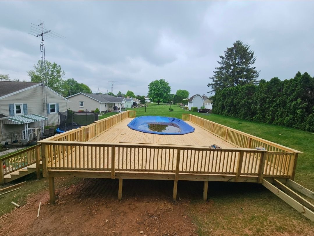 Wooden deck surrounding an above-ground pool with blue cover, in a grassy backyard, cloudy sky.