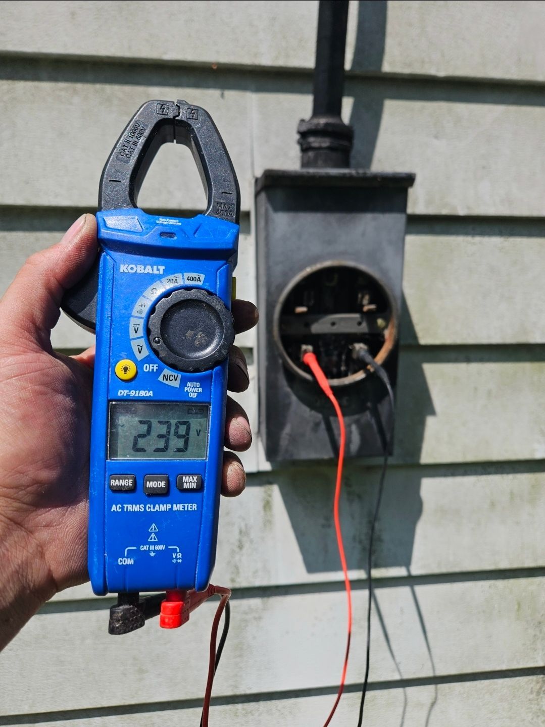 Hand holding a blue clamp meter reading 239 near a meter box with exposed wires.