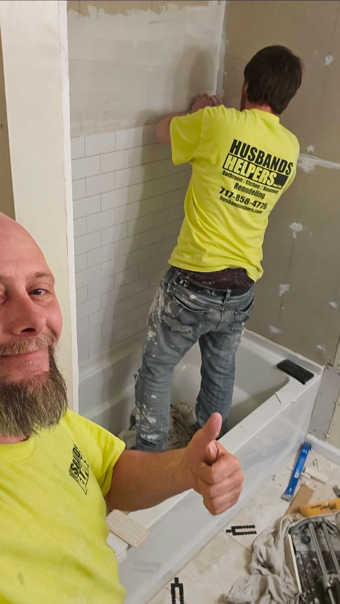 Two people tiling a shower, one giving a thumbs-up. Yellow shirts, paint-splattered jeans, setting.