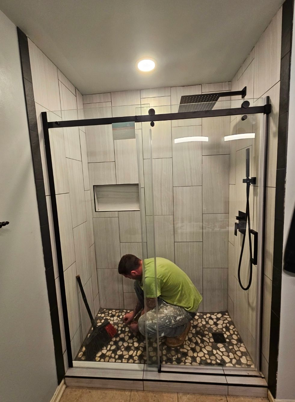 Man installing pebbles in a shower, next to glass door with black trim. White tile walls.
