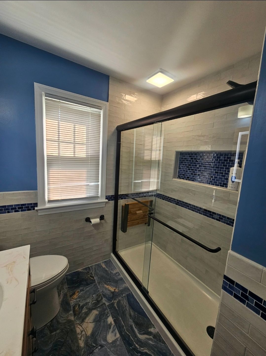 Blue and gray bathroom with shower, toilet, and window with blinds.