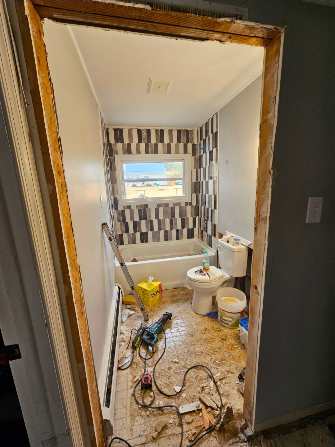 Bathroom remodel in progress: doorway view, exposed studs, debris on floor, patterned tile wall, toilet and tub.