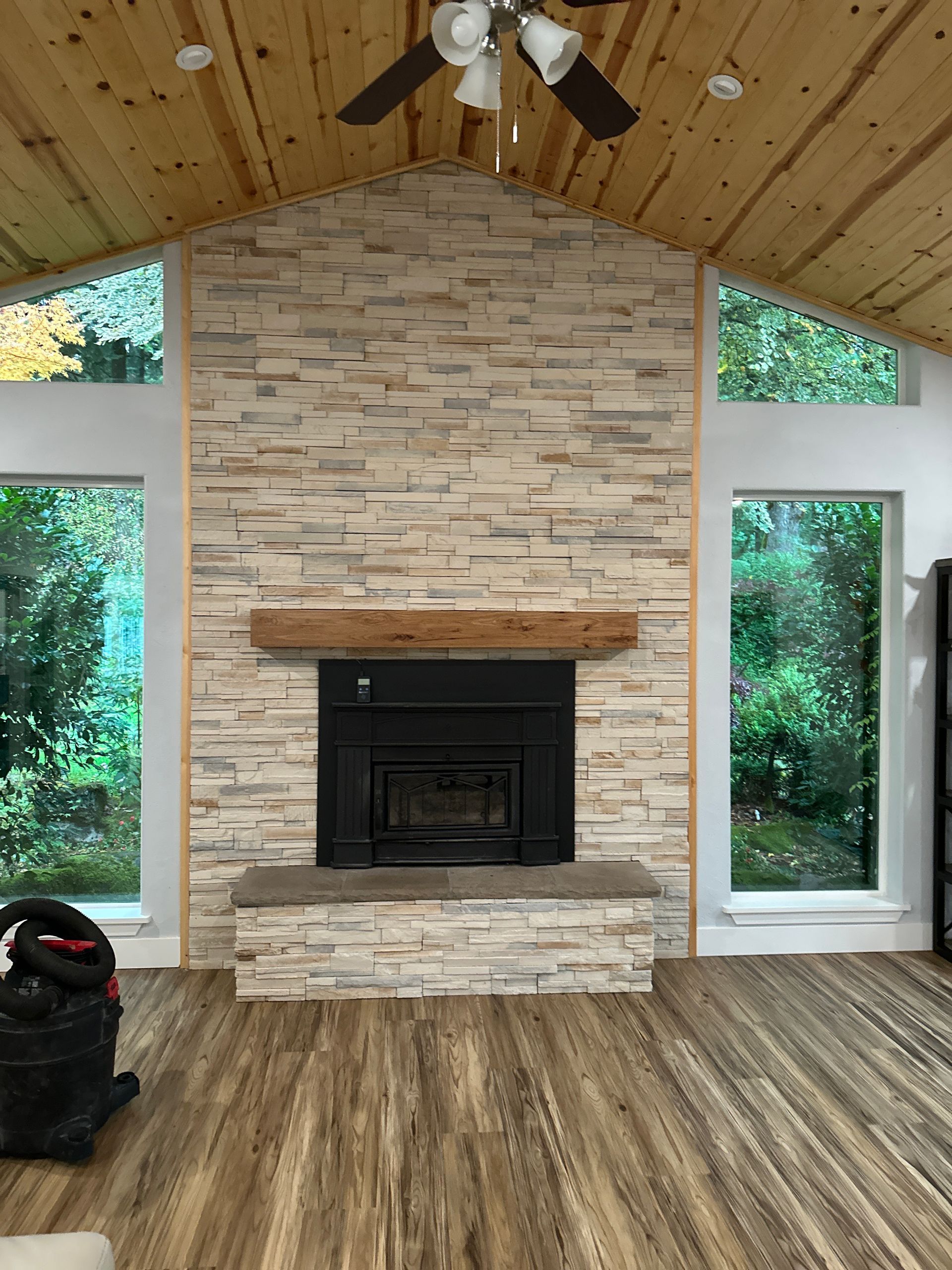 Fireplace with stone facade, wood mantel, and windows overlooking a lush green exterior. Wooden ceiling and floor.