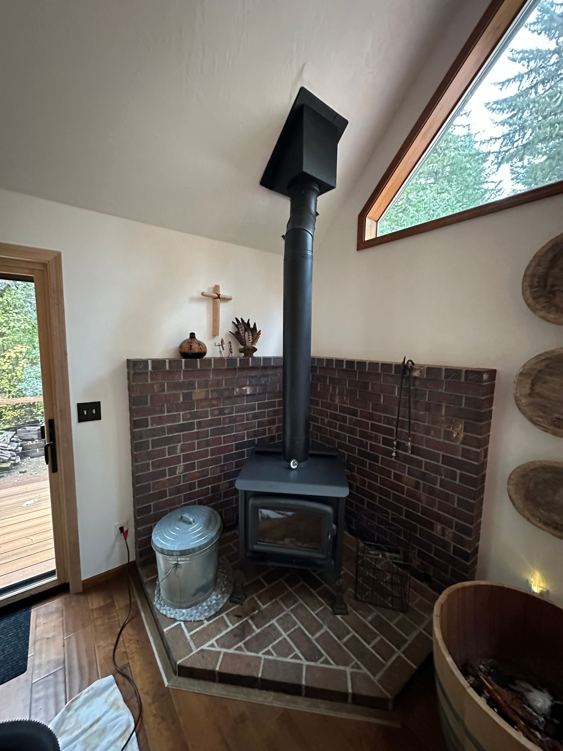 Wood-burning stove in corner brick enclosure, with black chimney. Small window with trees visible.