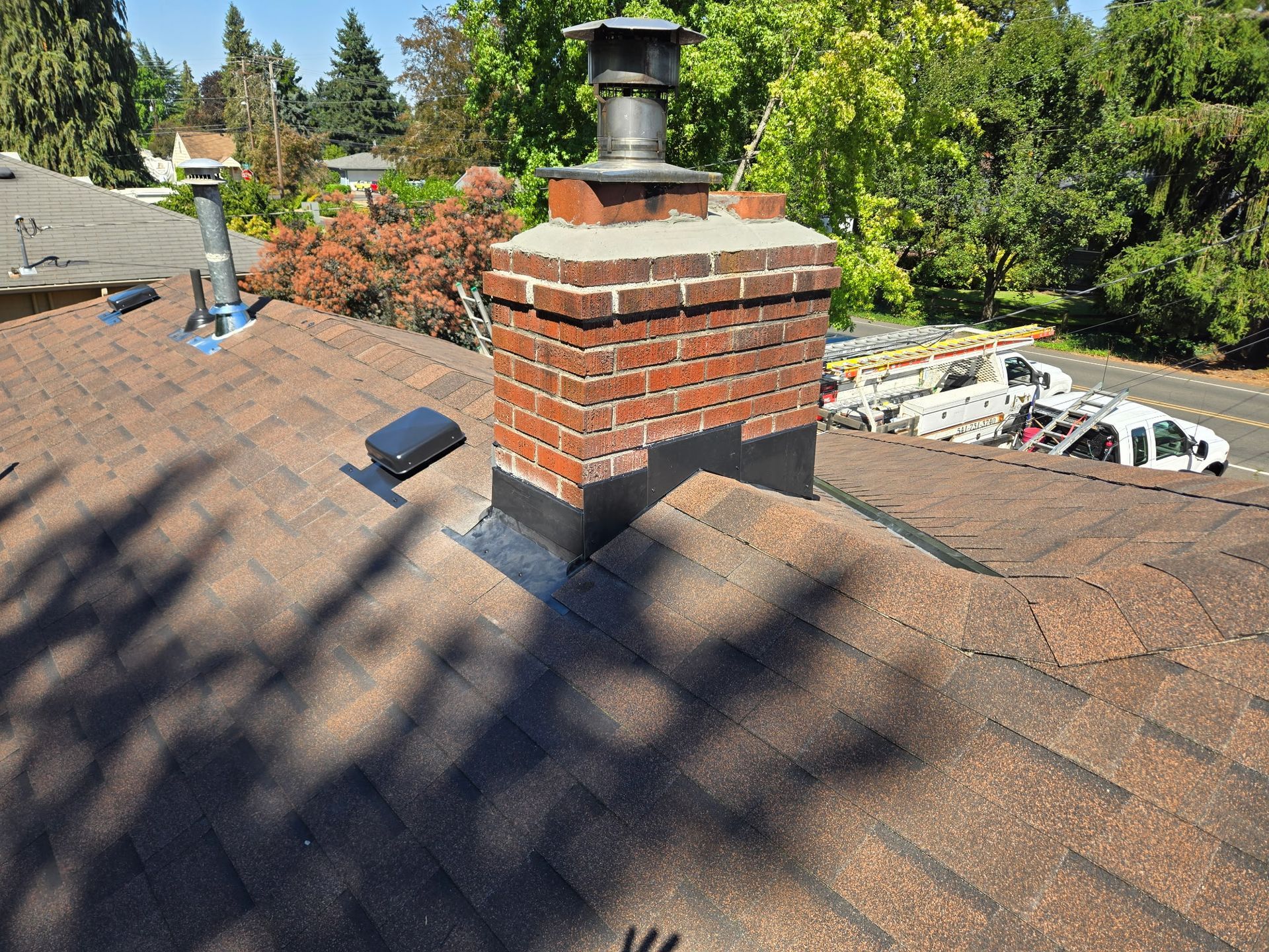 Brick chimney on a brown shingled roof, set against a sunny sky with trees and a street in the background.