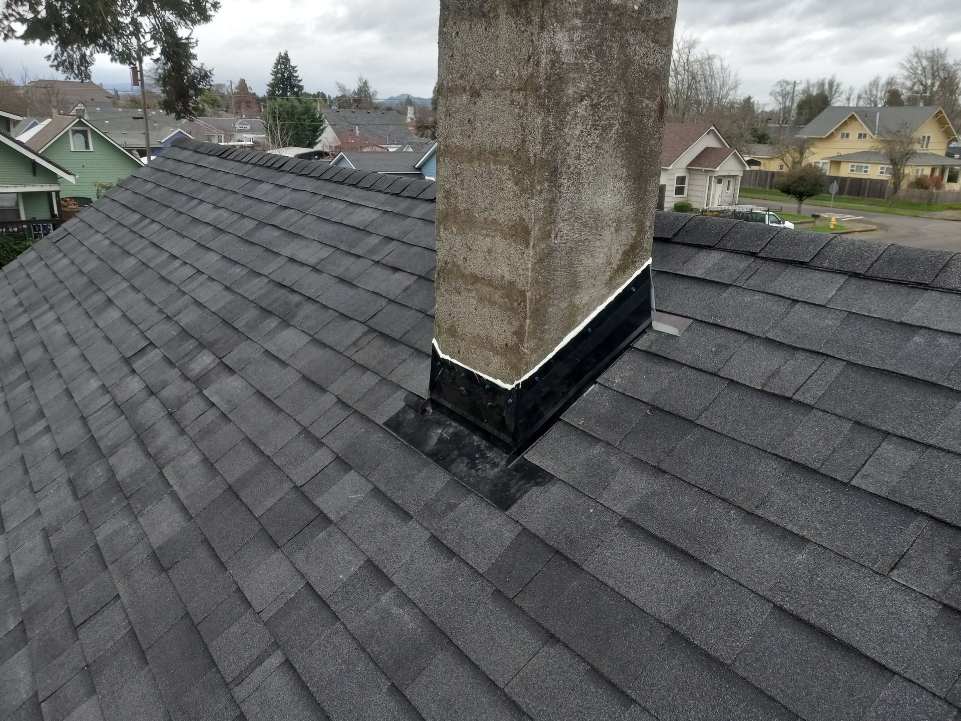 Gray asphalt shingle roof with a brick chimney. Flashing visible at chimney base. Cloudy sky in the background.