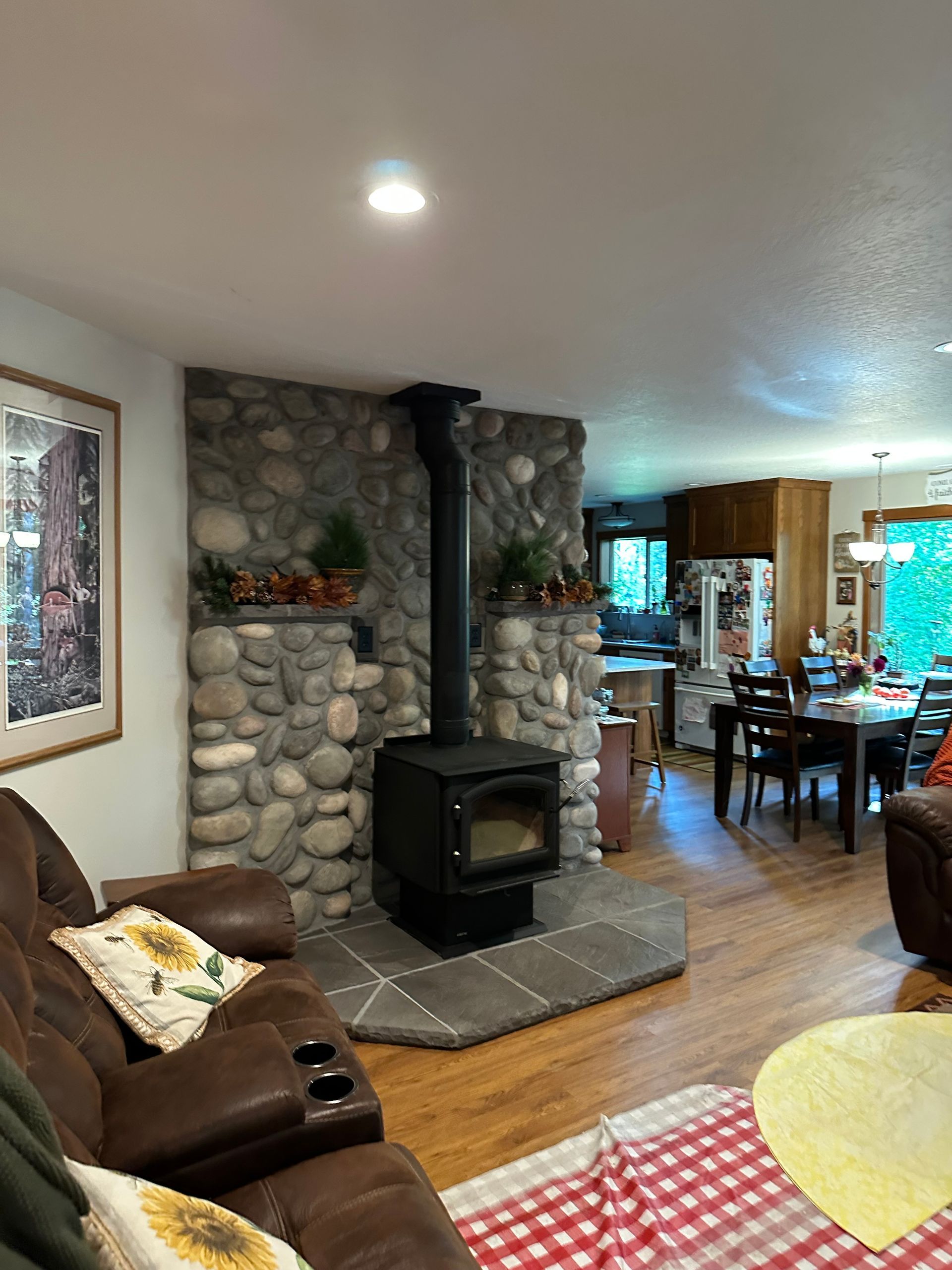 Cozy living room with stone fireplace, wood stove, and brown leather furniture. Kitchen and dining area visible.