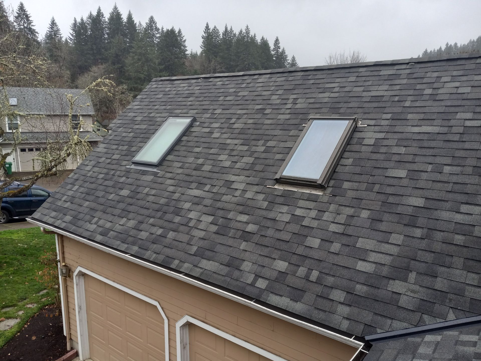 Dark gray shingle roof with two skylights, on a tan garage with trees in the background.