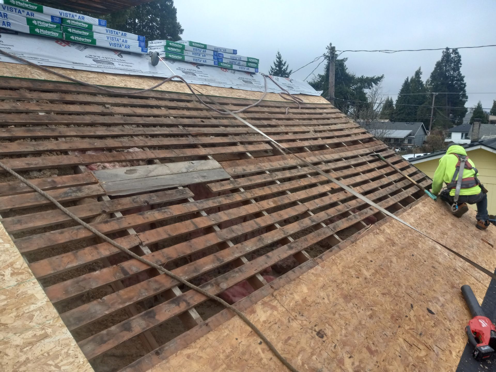 Roofer in safety gear on a roof, removing old slats. New OSB sheathing is installed.