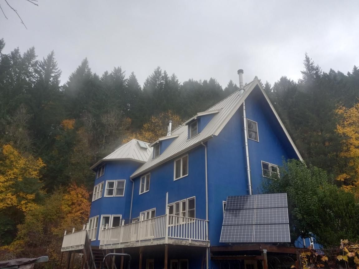 Blue house with white trim, deck, and windows against a fall forest backdrop under a cloudy sky.