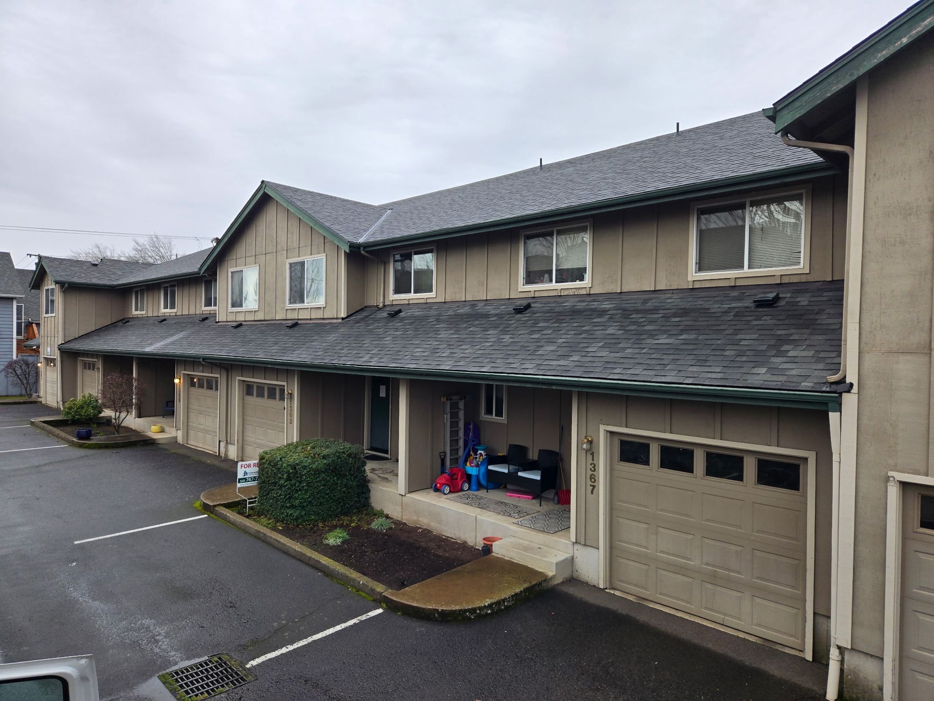 Row of townhouses with gray shingles and tan siding. Garage doors below. Overcast sky.
