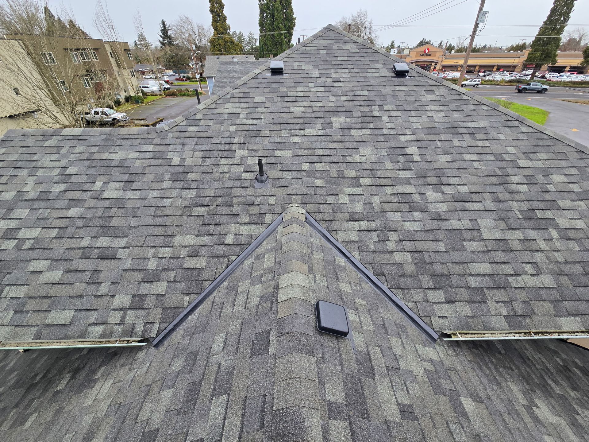 Gray shingle roof, view from above. Black trim and vents. Buildings and street visible in the background.