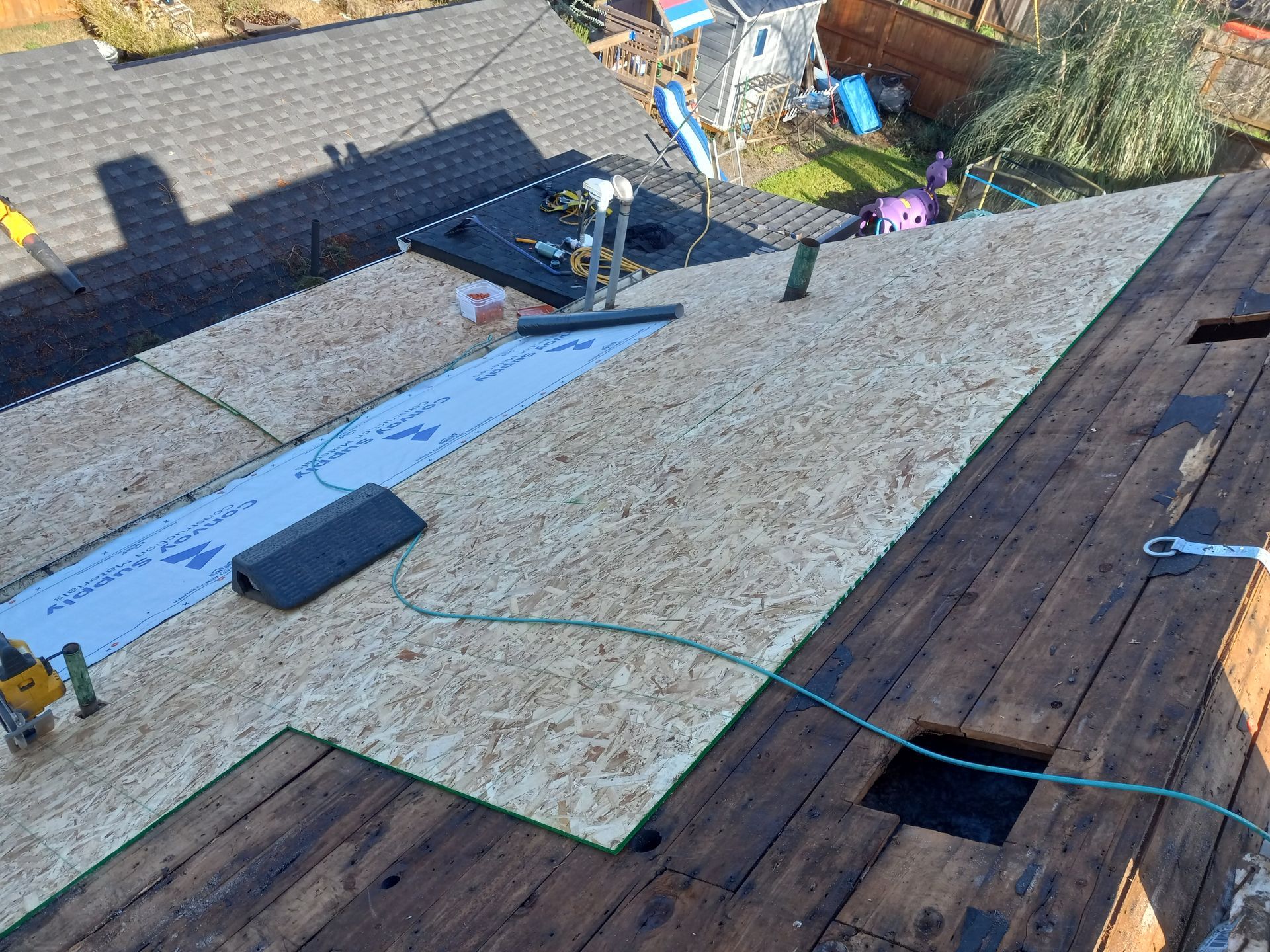 Rooftop with new wood sheathing being installed. Tools, safety line, and existing roof visible.