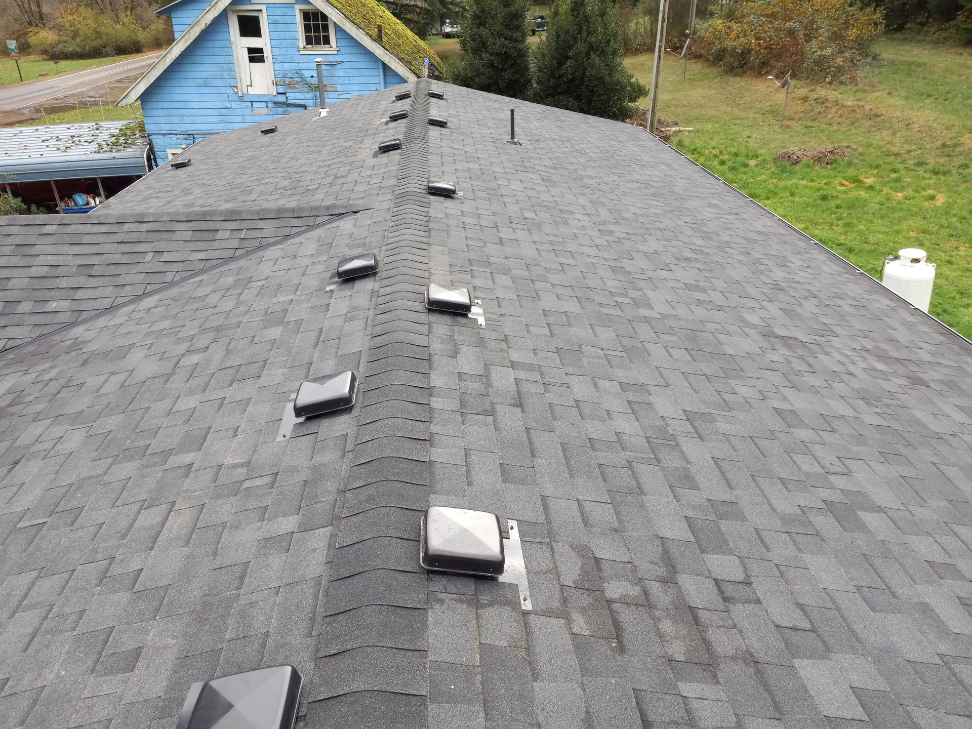 Roof with dark gray shingles and rooftop vents, viewed from the ridge toward a blue house in the background.