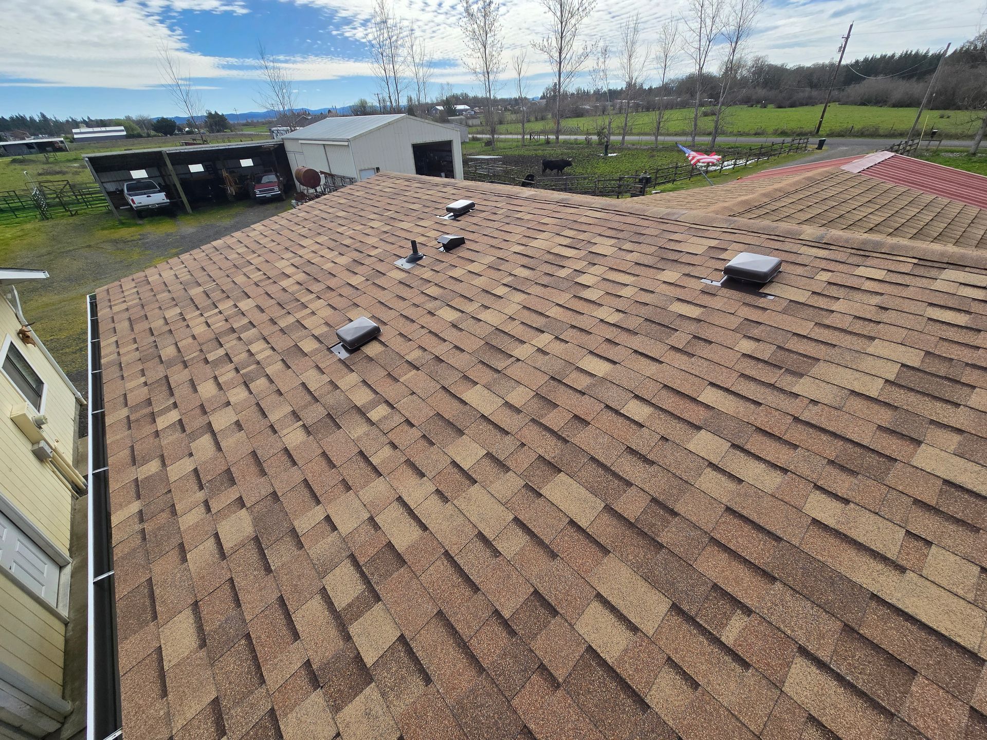 Rooftop with brown shingles, vents, and a wide view of fields under a blue sky.