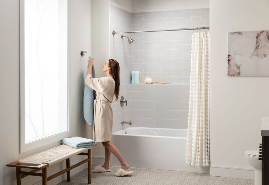 Woman hanging a towel in a modern bathroom with a window, shower, bench, and beige robe.
